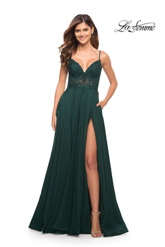 A woman smiles in a dark green La Femme Long A-Line Prom Dress with sheer bodice, thigh-high slit, spaghetti straps, and pockets. The "La Femme" logo is in the top right corner.