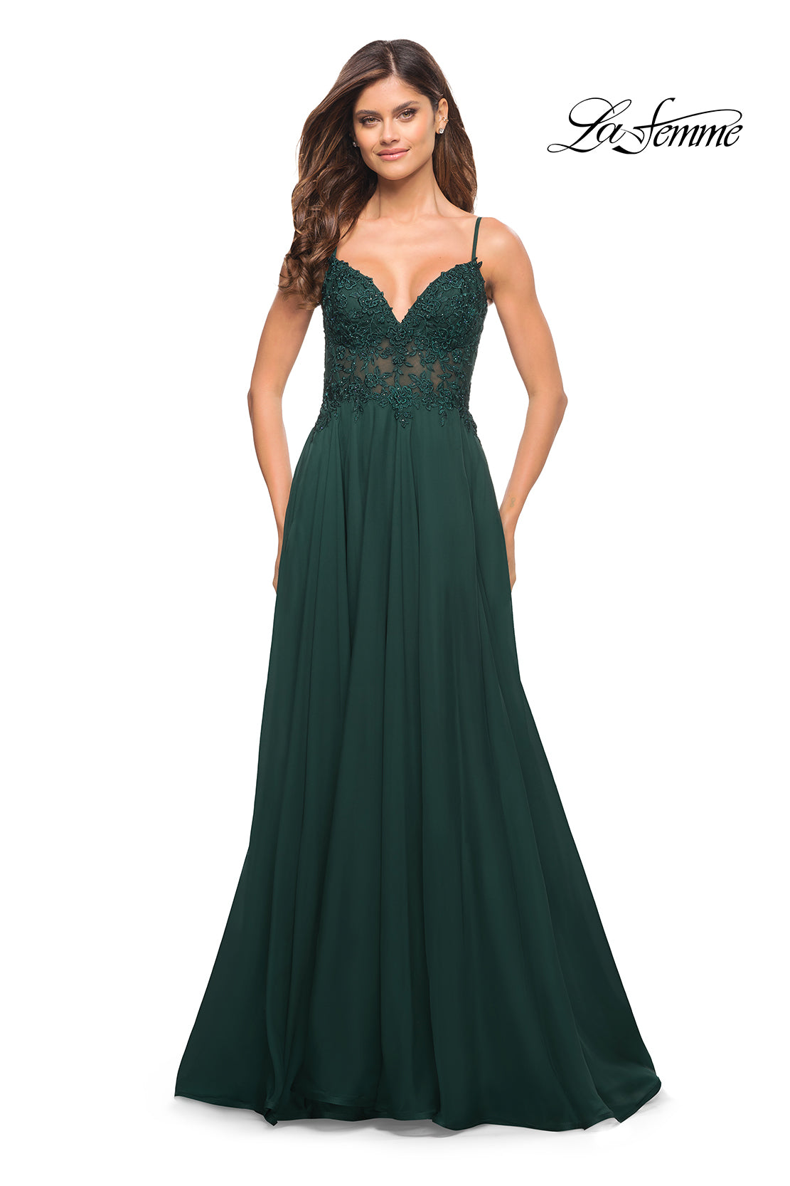 A woman wears the La Femme Long A-Line Prom Dress with a dark green flowing skirt, sheer lace bodice, and thin straps. The La Femme logo is displayed in the upper right corner.