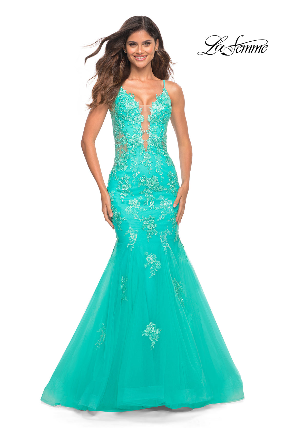 A woman wears the Aqua Blue Long Lace Prom Dress: La Femme 30675, featuring floral embroidery, a sheer bodice, thin straps, and a mermaid silhouette, standing against a white background.