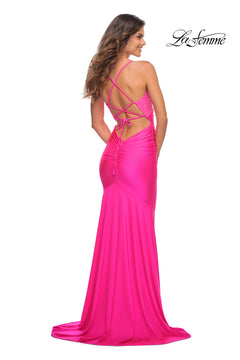 A woman stands looking over her shoulder in the Neon Pink Jersey La Femme Dress 30688—floor-length, backless, with crisscross straps and ruched detail. The "La Femme" logo appears in the top right corner.