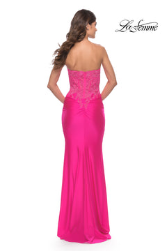A woman stands with her back to the camera, wearing the La Femme Strapless Neon Pink Prom Dress featuring lace detailing on the bodice.