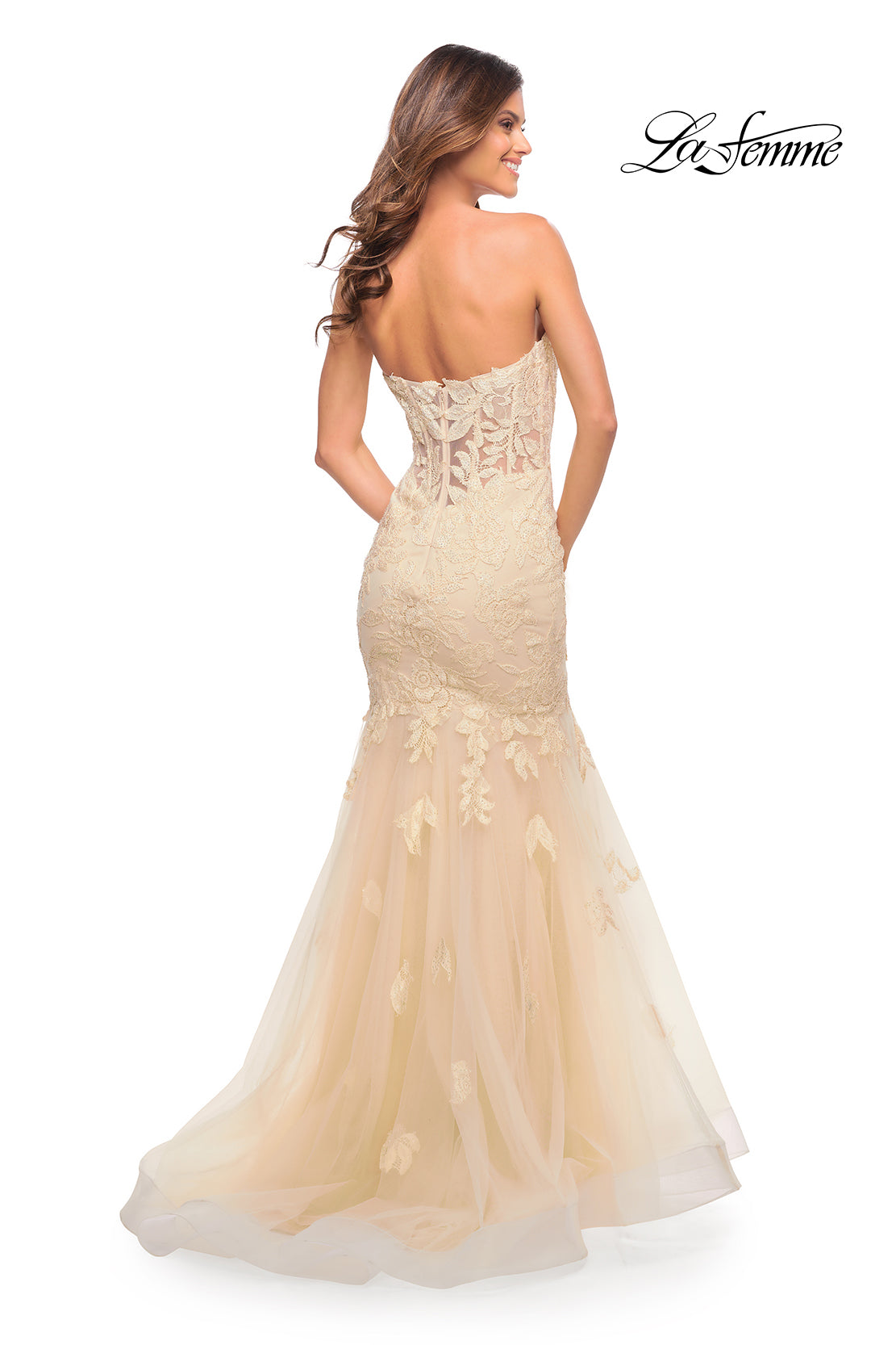 A woman faces away in a Sheer-Back La Femme Strapless Mermaid Prom Dress featuring a corset back, floor-length cream and nude lace overlay, and tulle skirt. 