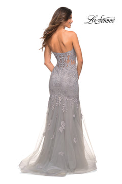 A woman models the Sheer-Back La Femme Strapless Mermaid Prom Dress in light gray. Photographed from the back, this floor-length gown features a lace corset back, floral appliqués, tulle accents, and a partially sheer bodice.