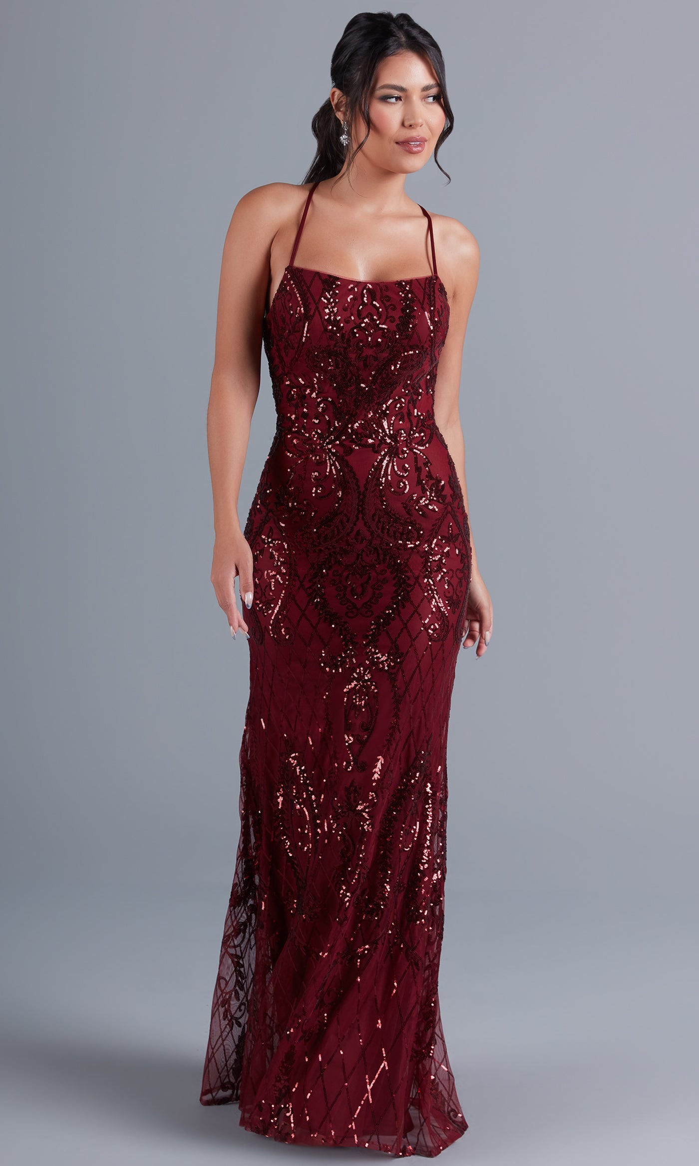A woman models the PromGirl Statement-Back Long Sequin Prom Dress, a sleeveless burgundy gown with thin straps and a square neckline, standing against a plain gray background.