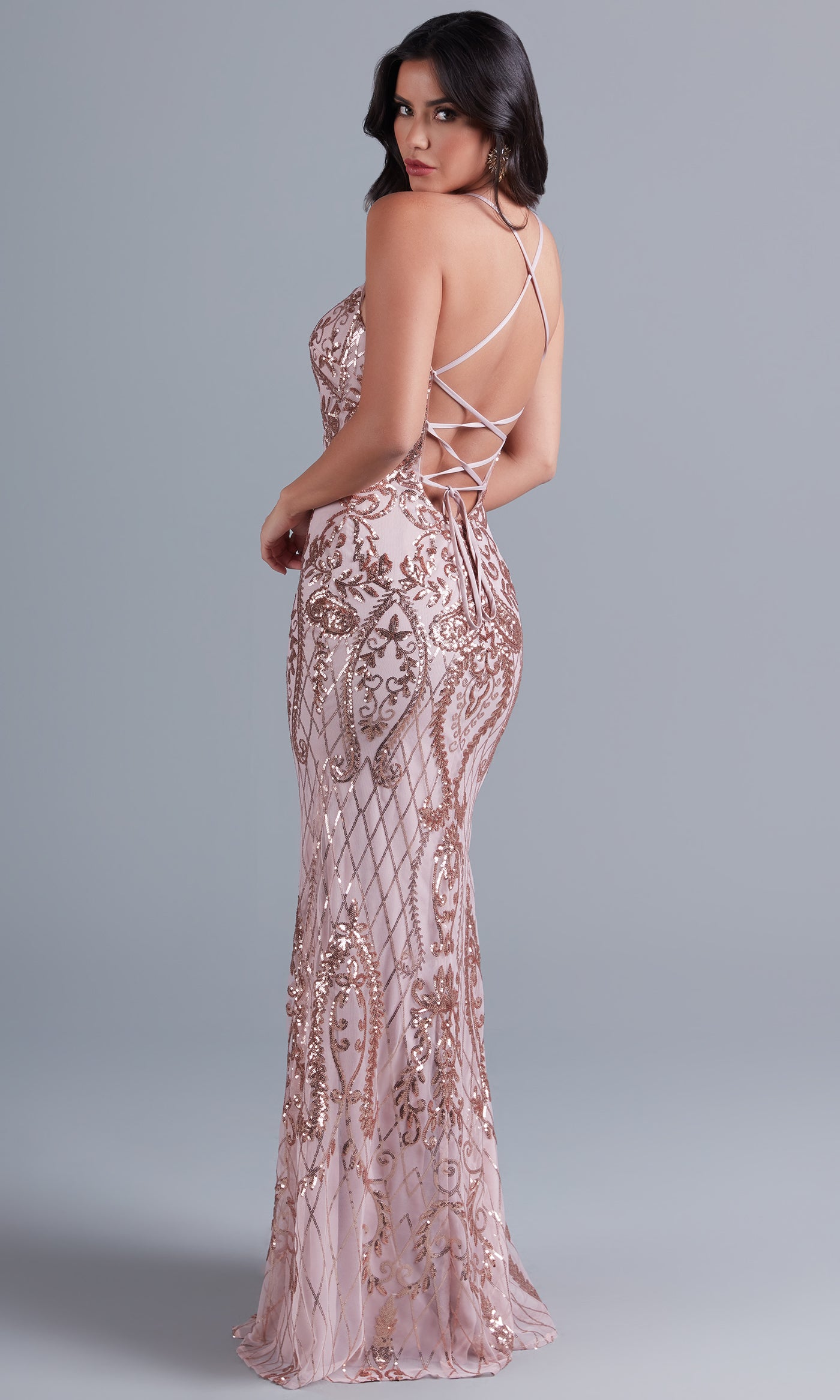 A woman poses in the PromGirl Statement-Back Long Sequin Prom Dress, featuring a blush pink hue and crisscross lace-up back straps for a stunning statement-back prom look.