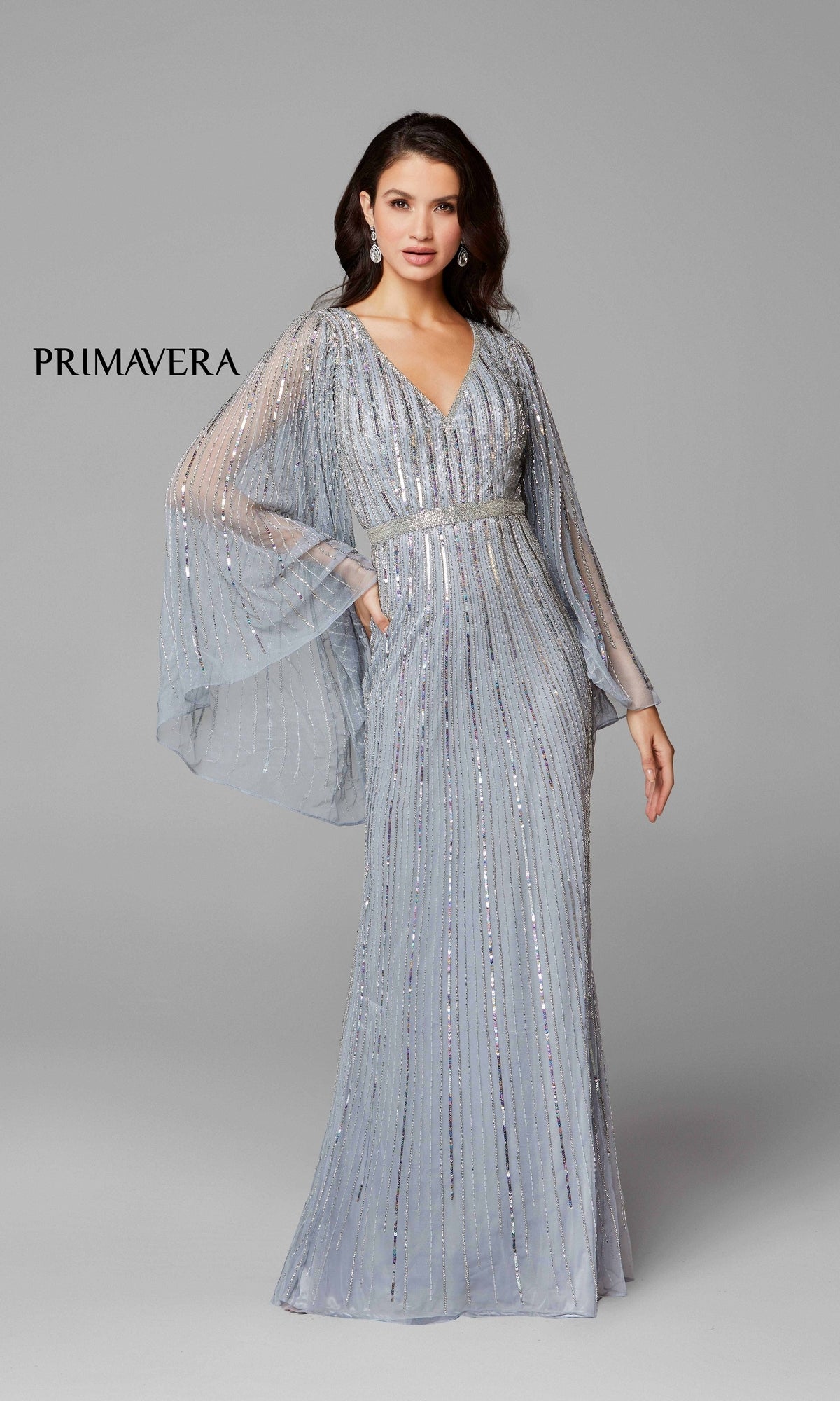 A woman models the Dramatic Sheer-Long-Sleeved Long Formal Dress 3672—silver, sequined, and floor-length with sheer sleeves—against a gray background. “PRIMAVERA” is displayed on the left.