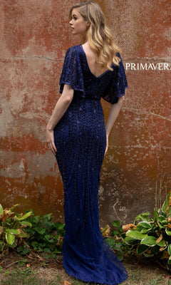 A woman wearing the Short-Sleeve Beaded Long Prom Dress 3681 in dark blue stands outdoors, facing away from the camera, before a weathered wall with greenery at its base.