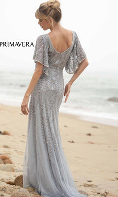 A woman stands on a sandy beach facing the ocean, wearing the Short-Sleeve Beaded Long Prom Dress 3681, its silver beading shimmering in the light as her hair is styled in an elegant updo.