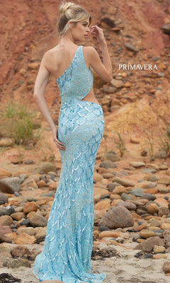 A woman stands on rocky terrain in the Primavera 3729 One-Shoulder Long Sequin Prom Dress, featuring a light blue hue, waist cutout detail, and shimmering sequins against a reddish-brown, sandy hillside.