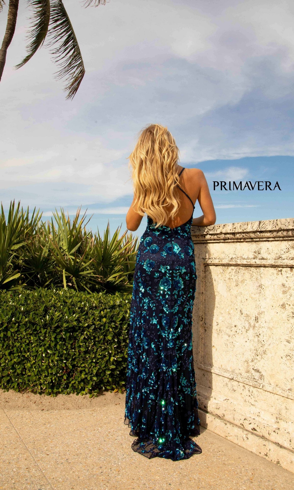 A woman with long blonde hair in a blue Primavera 3731 Sequin-Print Long Formal Prom Dress stands outside by a stone wall and greenery, facing away. The sky is partly cloudy, and 