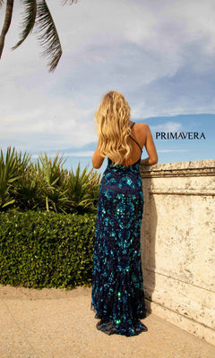 A woman with long blonde hair in a blue Primavera 3731 Sequin-Print Long Formal Prom Dress stands outside by a stone wall and greenery, facing away. The sky is partly cloudy, and "PRIMAVERA" appears on the wall.