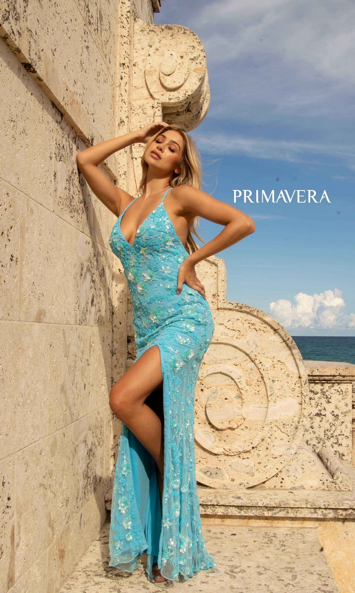 A woman poses by a stone wall near the ocean in the Primavera 3731 Sequin-Print Long Formal Prom Dress, featuring a light blue color and thigh-high slit; 