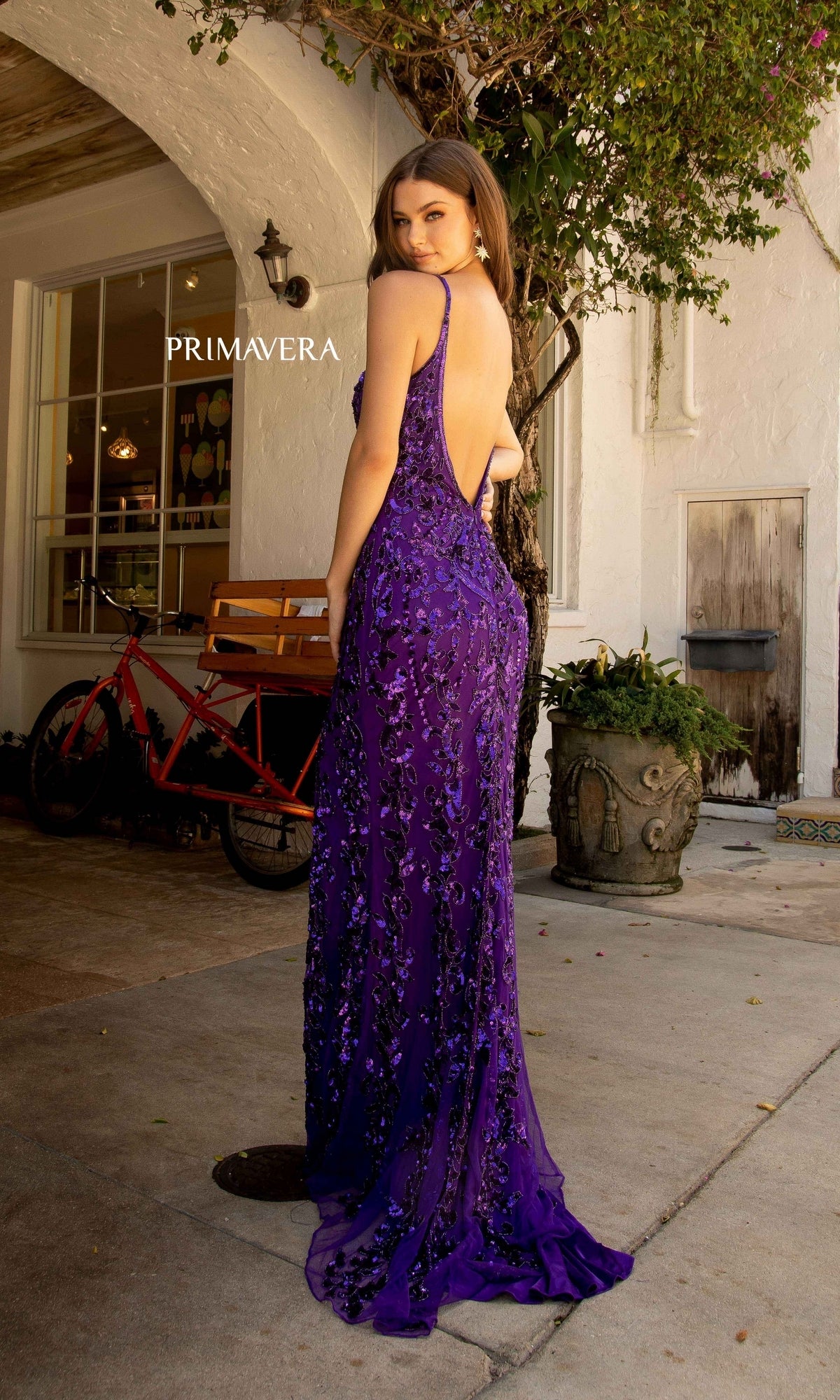A woman stands outdoors in the Open-Back Primavera 3749 Long Sequin Prom Dress—a sleeveless, purple evening gown with sparkling details—glancing over her shoulder, as a red bicycle and lush greenery enhance the elegant setting.