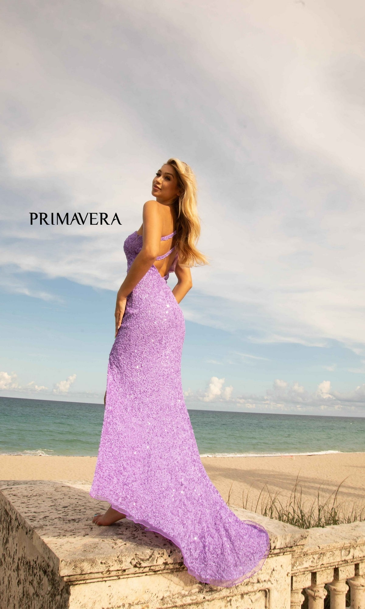 A woman wearing the Bright Neon Primavera One-Shoulder Prom Dress 3761N stands on a stone ledge by the beach, gazing at the ocean beneath a partly cloudy sky. 
