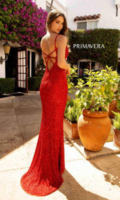 A woman in the Primavera Dark Sequin Long Prom Dress 3792D, featuring a sparkling red design and strappy open back, stands outdoors near potted plants, glancing over her shoulder with "PRIMAVERA" visible in the background.
