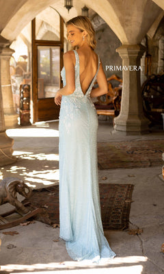 A woman wears the Primavera 3923 Backless Long Beaded Prom Gown, standing in an arched stone hallway decorated with rugs and antique pieces.