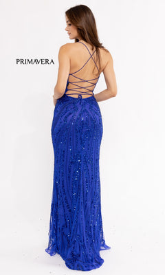 A woman faces away, wearing the Strappy-Back Long Sequin Prom Dress 3958 in royal blue. The elegant, floor-length gown features a low back with crisscross straps and sparkling sequins. "PRIMAVERA" is displayed on the left side.