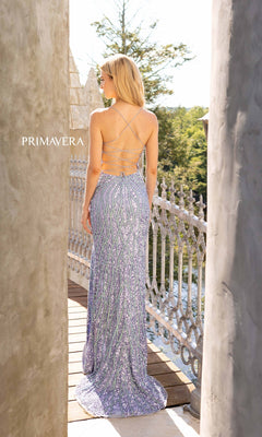 A woman with blonde hair stands on a balcony in the Long Beaded Primavera Prom Gown 3959, a lavender, sequin-covered backless dress with crisscross straps. Trees and a river create a serene backdrop for her elegant look.