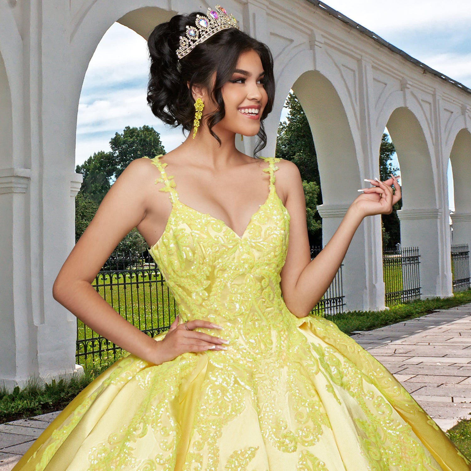 A young woman wearing the Quinceañera Sample Dress QF106, a yellow ornate v-neck gown with a tiara, stands outdoors by a white arched building, smiling and raising one hand.