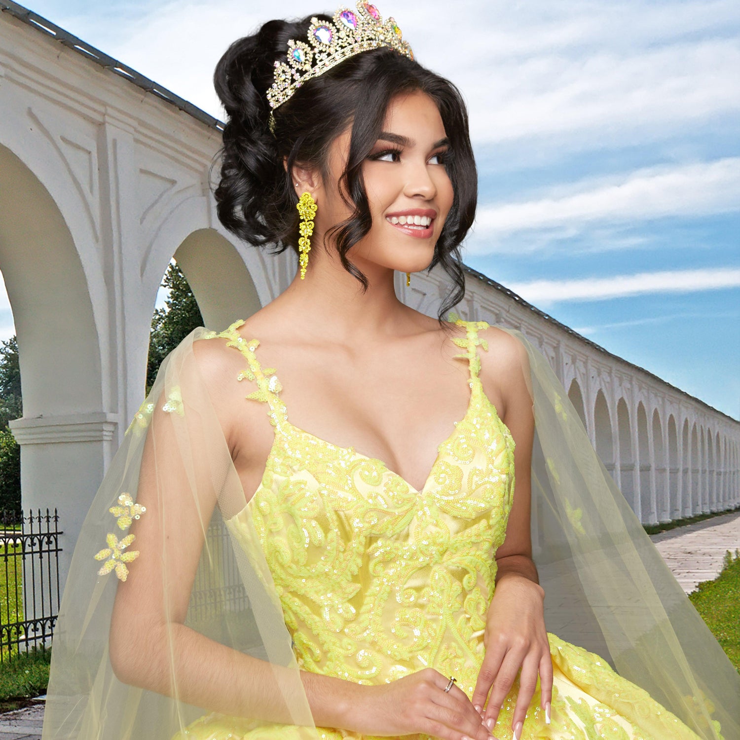 A young woman smiles and poses near a white arched colonnade outdoors, wearing the elegant Quinceañera Sample Dress QF106 and a tiara.