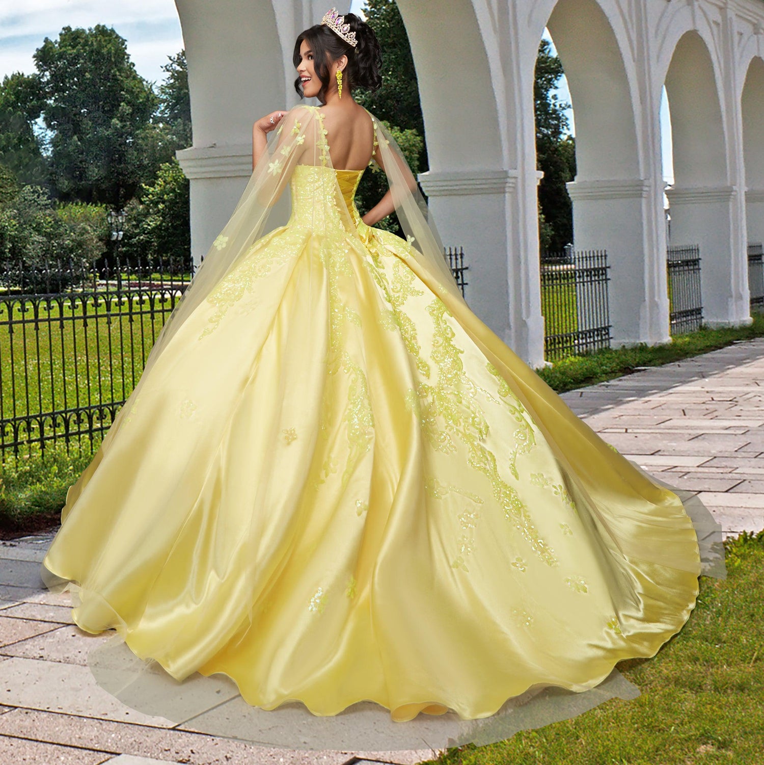 A woman stands outdoors on a stone path near a white arched building and black iron fence, facing away and wearing the shimmering Quinceañera Sample Dress QF106 in yellow with a v-neck, tiara, and elegant ball gown style.