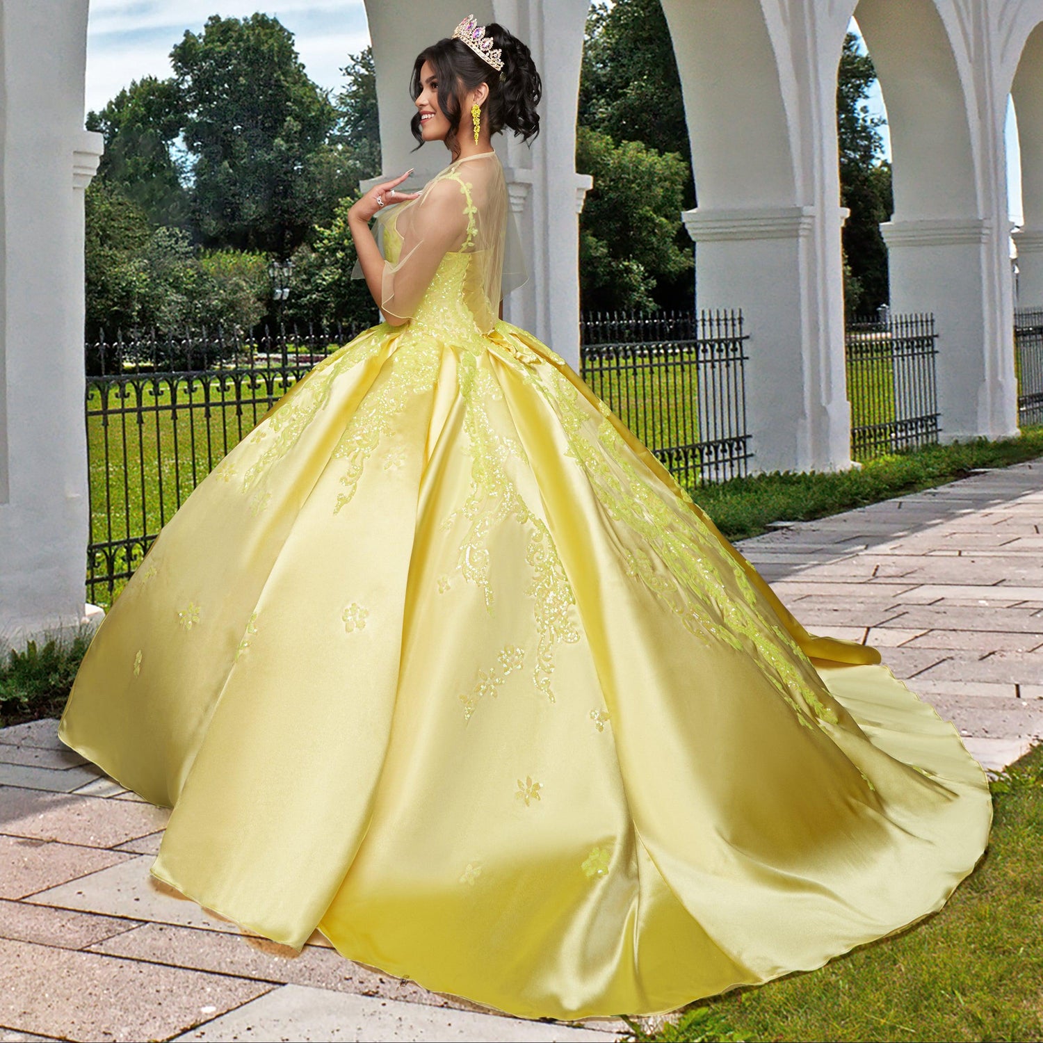 A young woman models the Quinceañera Sample Dress QF106, a yellow v-neck gown, and a tiara while standing outdoors near white arches and a black iron fence with greenery in the background.