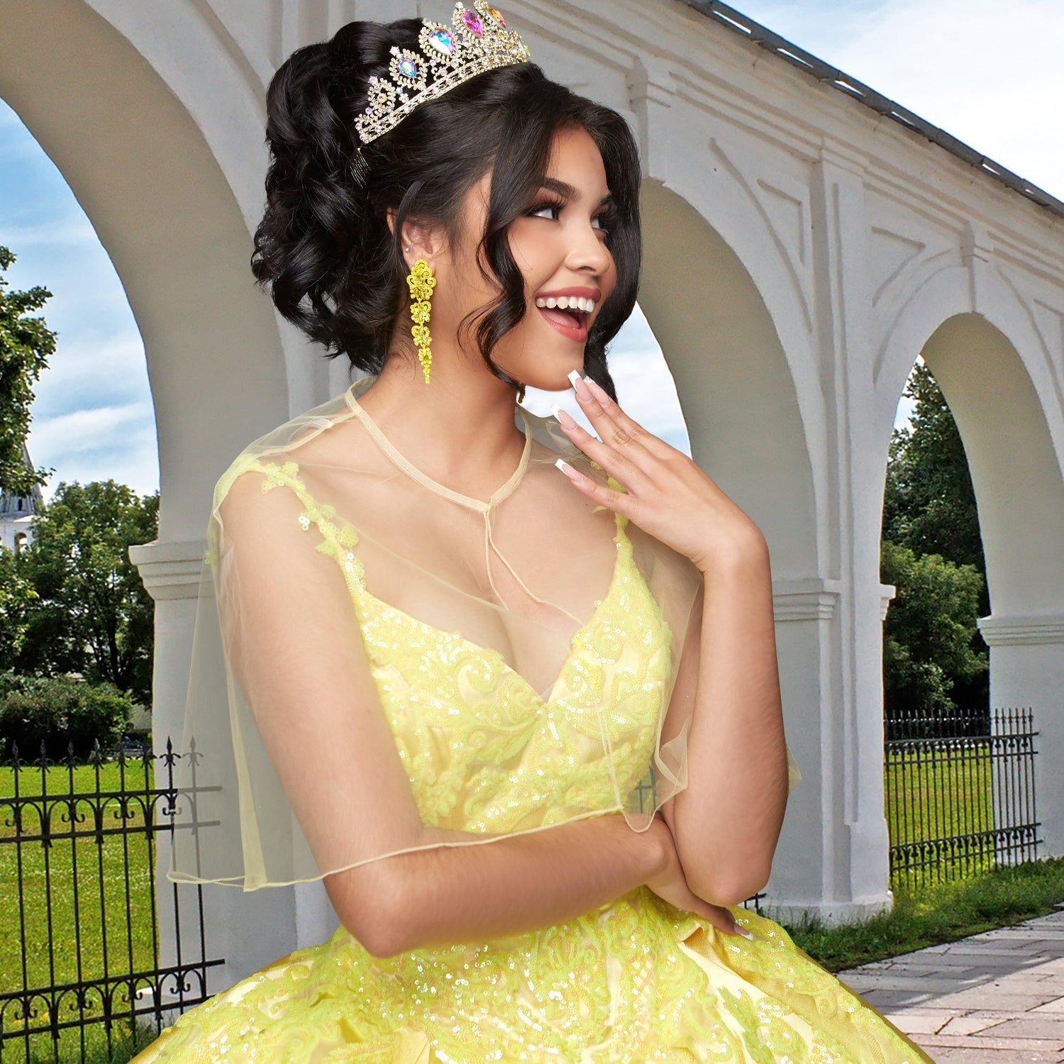A young woman stands outdoors, smiling with one hand near her face, wearing the Quinceañera Sample Dress QF106—a yellow sequin gown and tiara—against a backdrop of white arches and greenery.