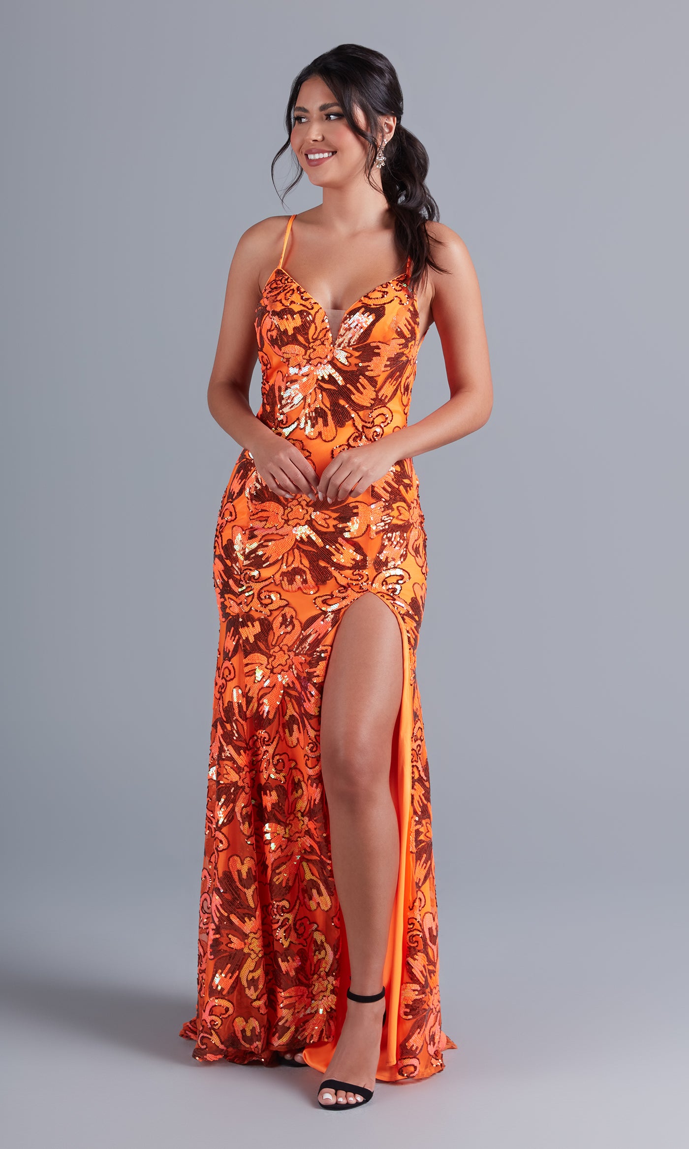 A woman models the Long Bright Sequin Unique Prom Dress RX5024—an orange, floor-length gown with thin straps, sequin details, and a high thigh slit—standing before a plain gray background.