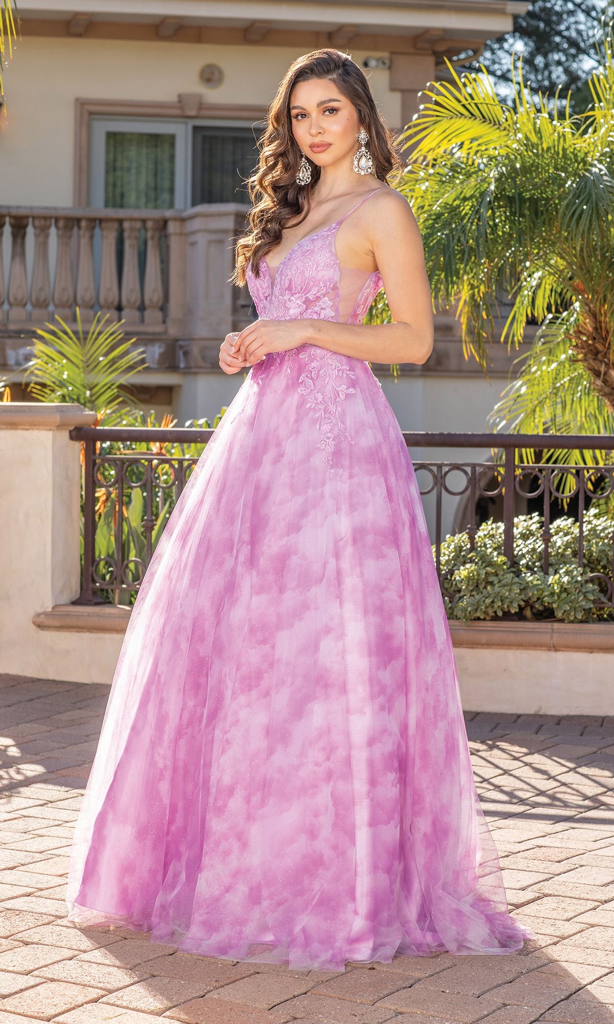 A woman in an affordable Tie-Dye Prom Ball Gown with Sheer Sides—sleeveless, floor-length, pink and purple—stands outdoors by a railing with palm trees and a building behind her.