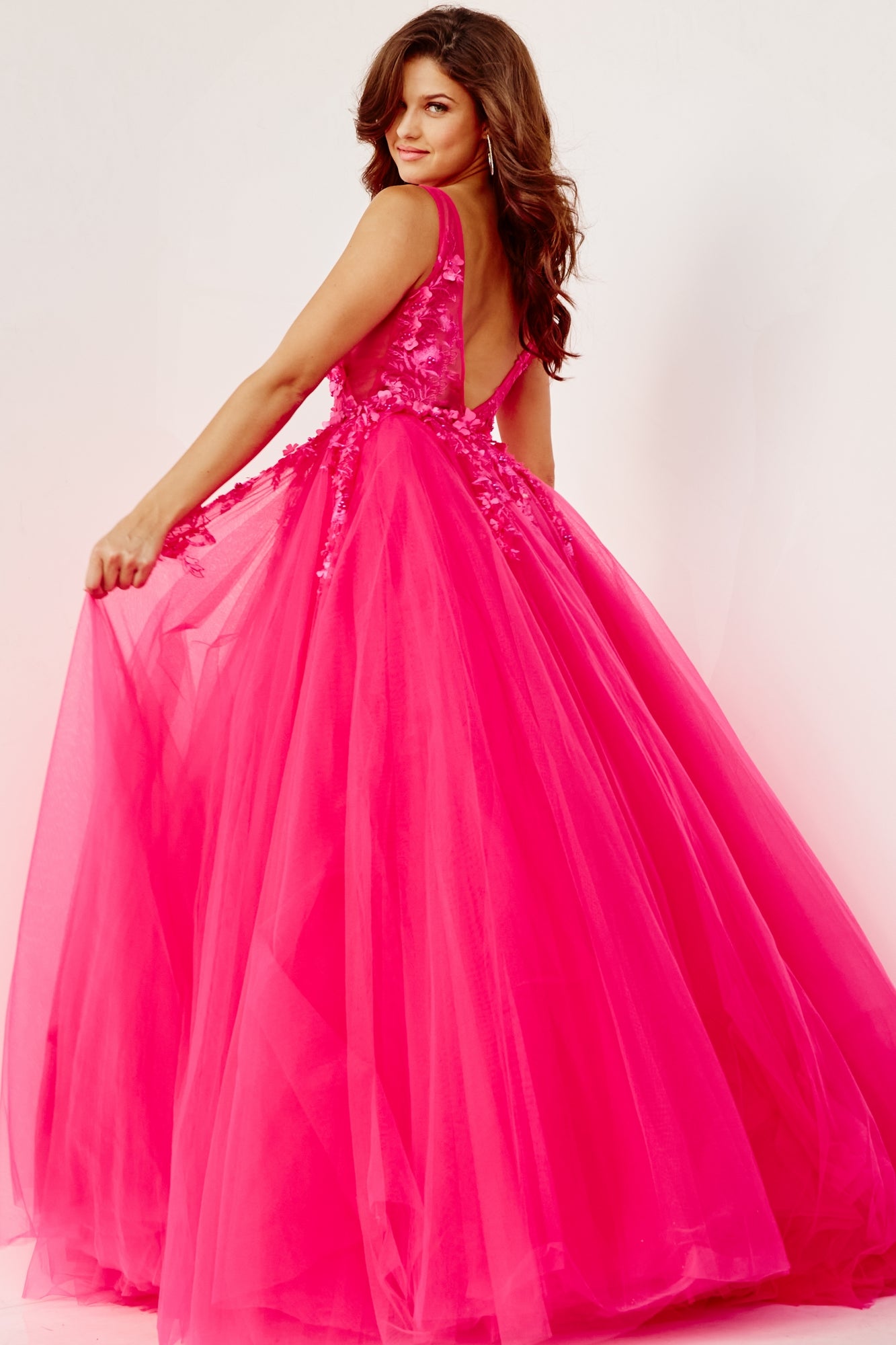 A woman in a bright pink Jovani Tulle Prom Ball Gown with floral appliques, featuring a floor-length ball gown silhouette and deep V back, stands against a plain background, looking over her shoulder while holding part of the dress.