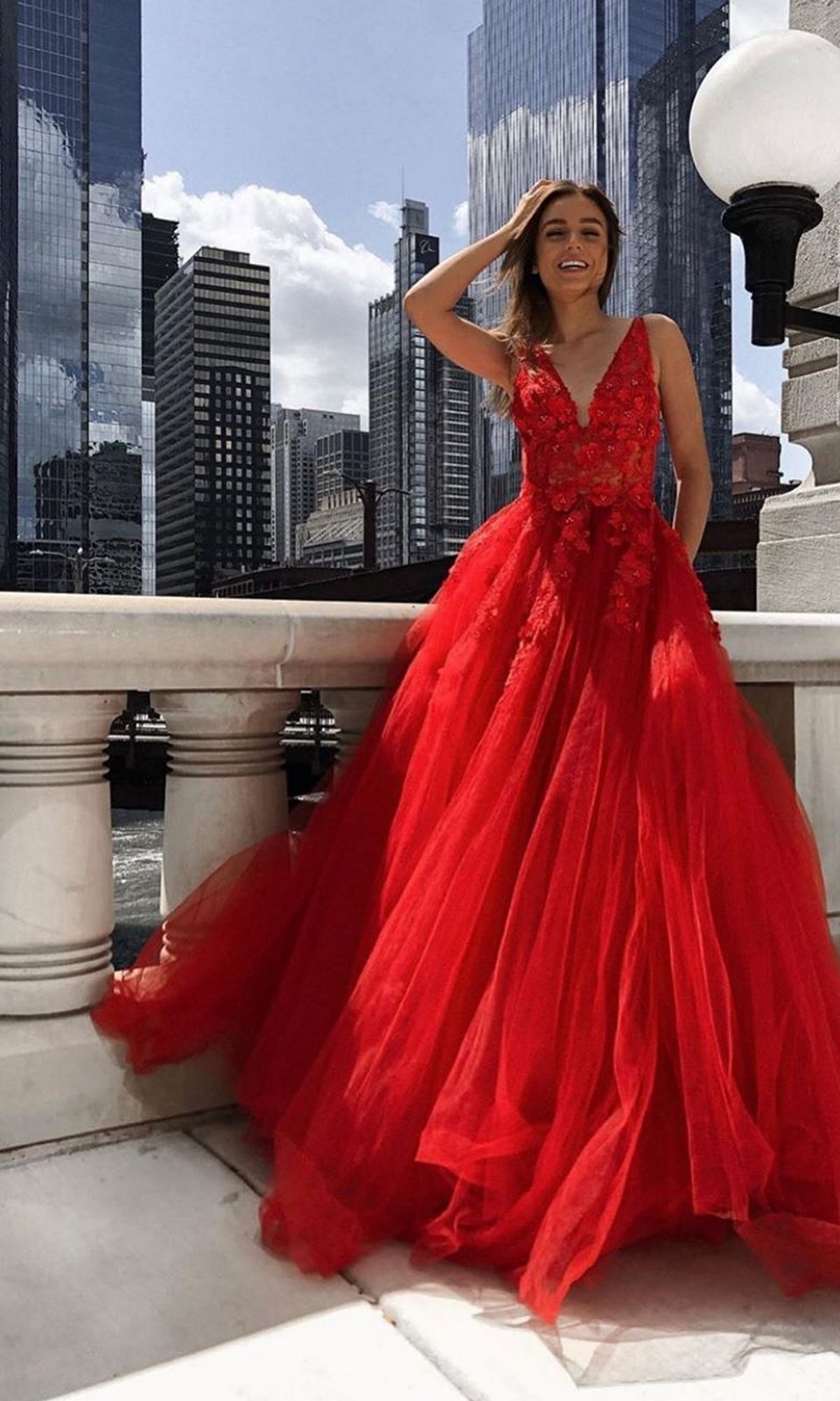 A woman in a Jovani Tulle Prom Ball Gown with Floral Appliques stands on a city balcony, skyscrapers behind her, smiling as she holds her hair.