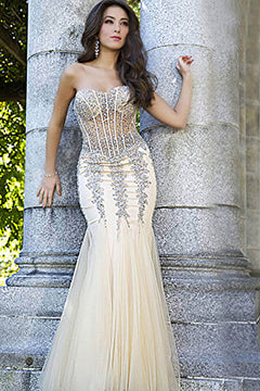 A woman wearing the Long Strapless Sweetheart Jovani Prom Dress 5908, featuring a fitted mermaid silhouette, sheer panels, and silver embellishments, stands by an outdoor stone pillar.