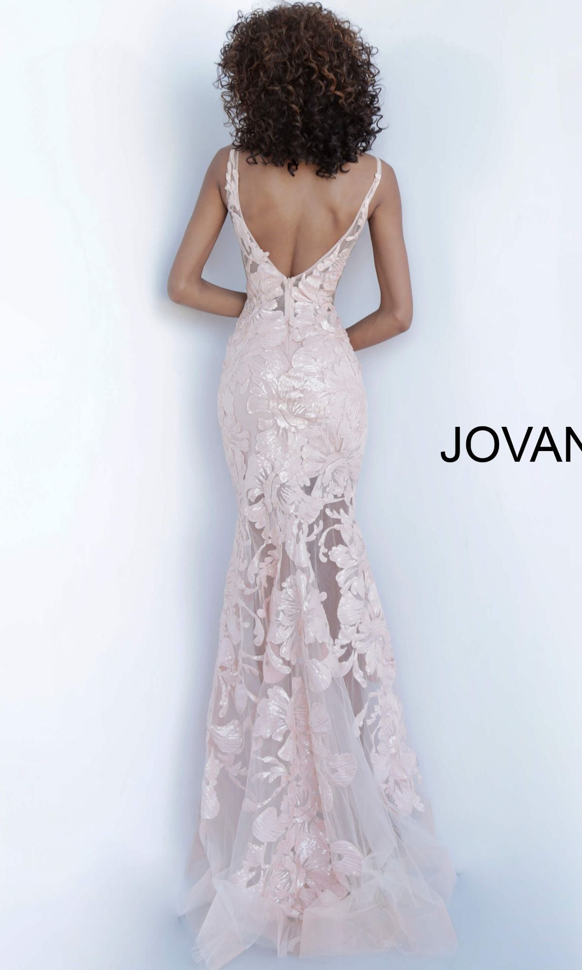 A woman with curly hair models the Jovani Embroidered Long Prom Dress with Sequins, a backless, blush pink, floor-length gown featuring floral lace embroidery, against a plain light background.
