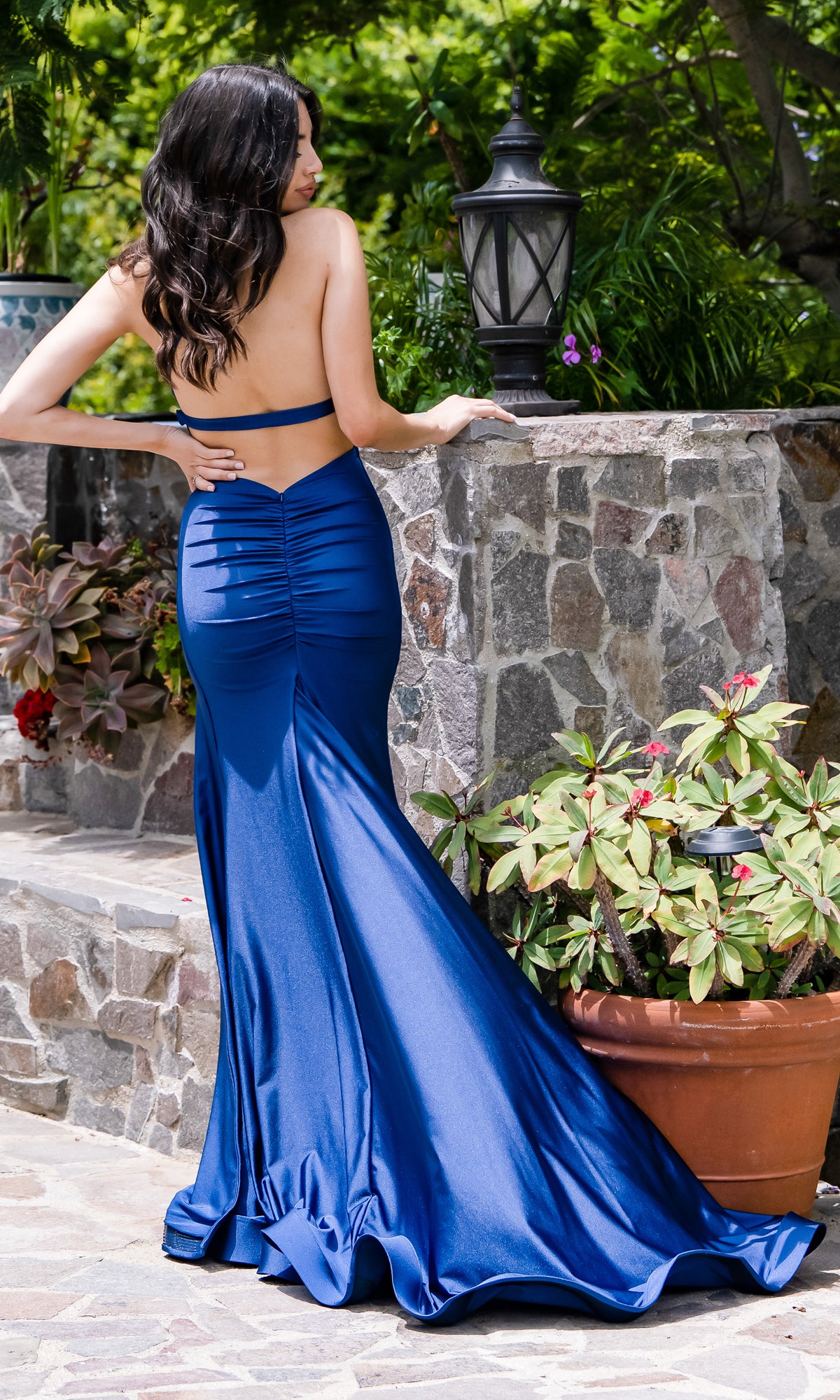 A woman stands outdoors by a stone wall and potted plants, facing away in the Atria One-Shoulder Sexy Long Prom Dress 6802H, featuring a fitted, backless royal blue design.