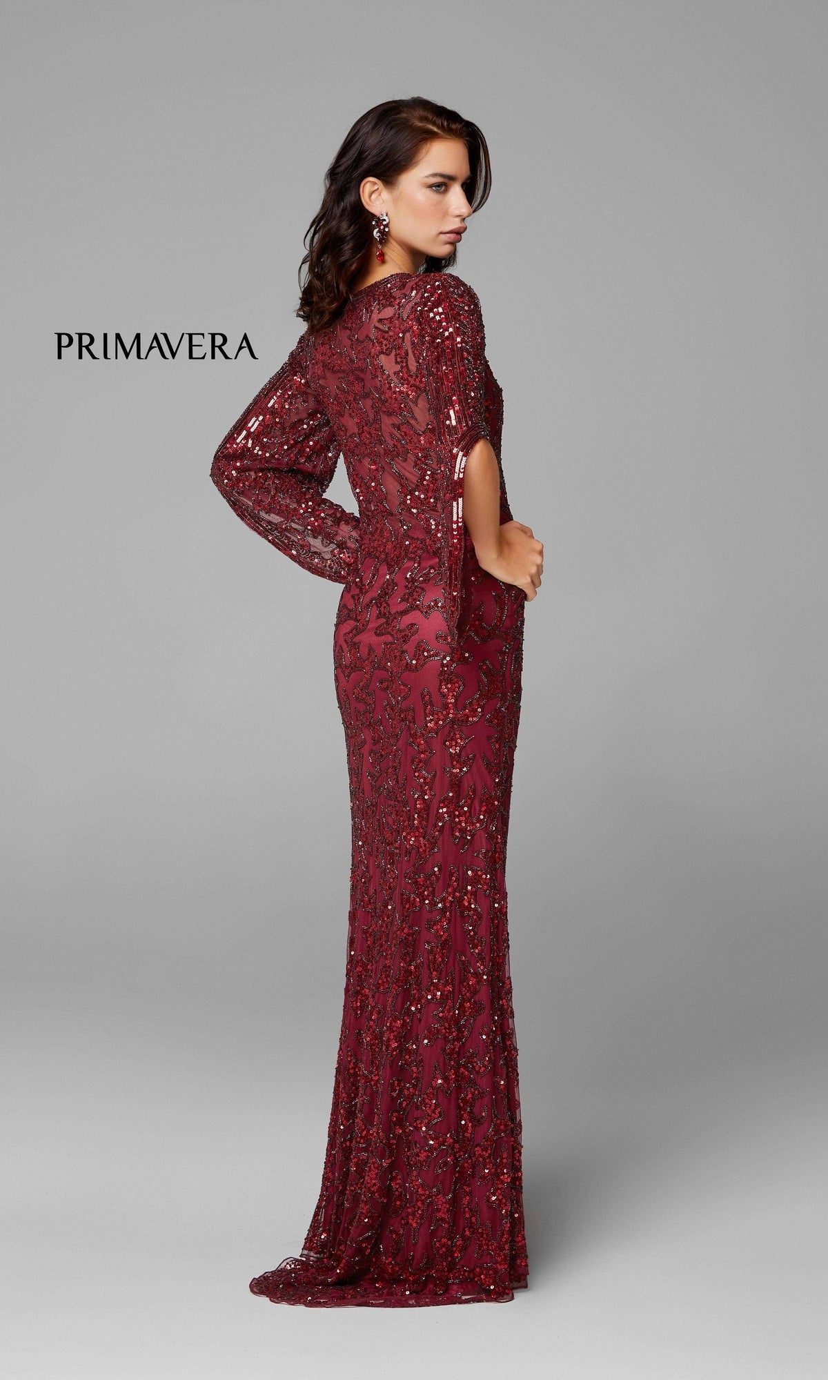 A woman in the Long Bell Sleeve Beaded Evening Gown 9713, featuring maroon sequins and sheer sleeves, stands sideways before a plain grey background. 