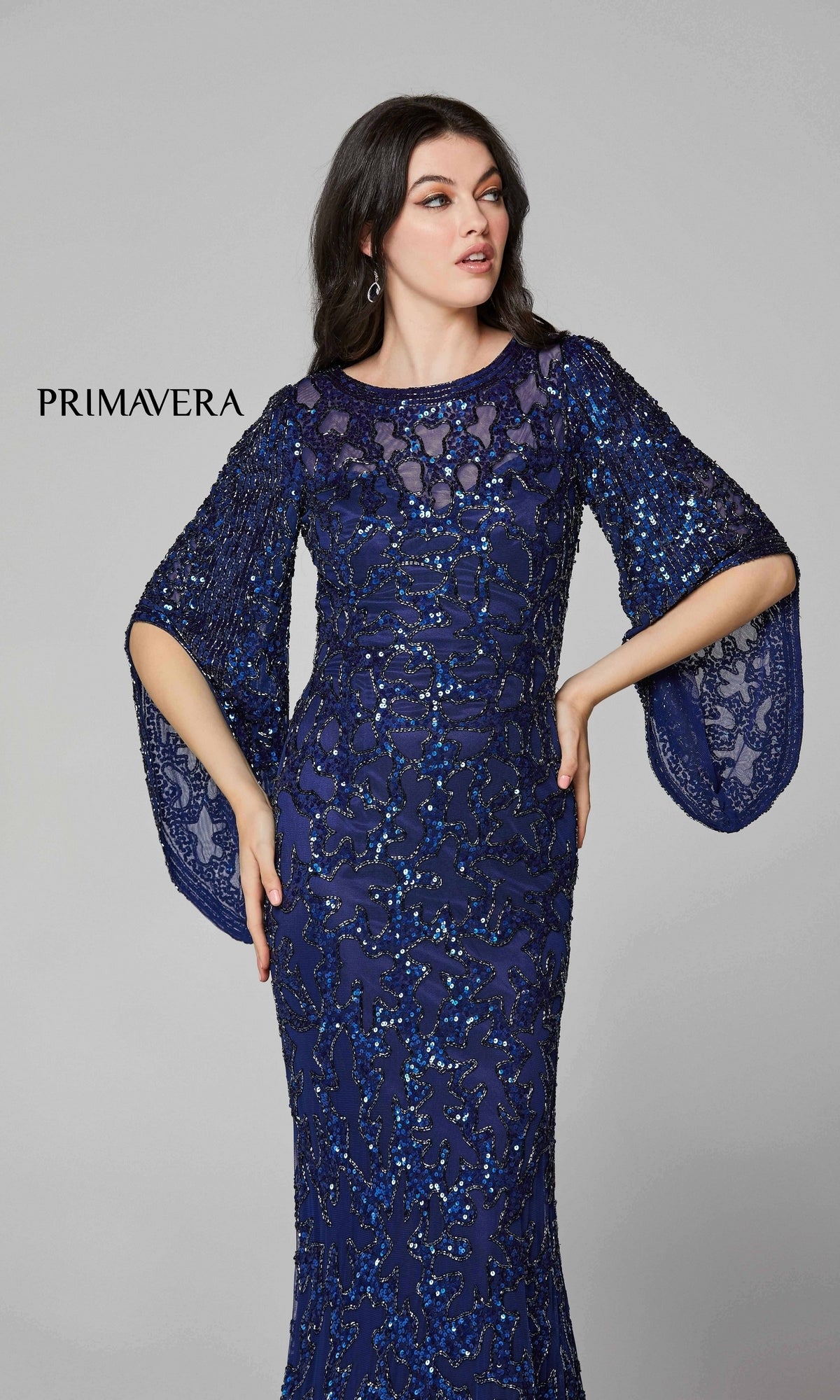 A woman models the Long Bell Sleeve Beaded Evening Gown 9713, a floor-length navy dress with sheer wide bell sleeves, against a plain light gray background. 