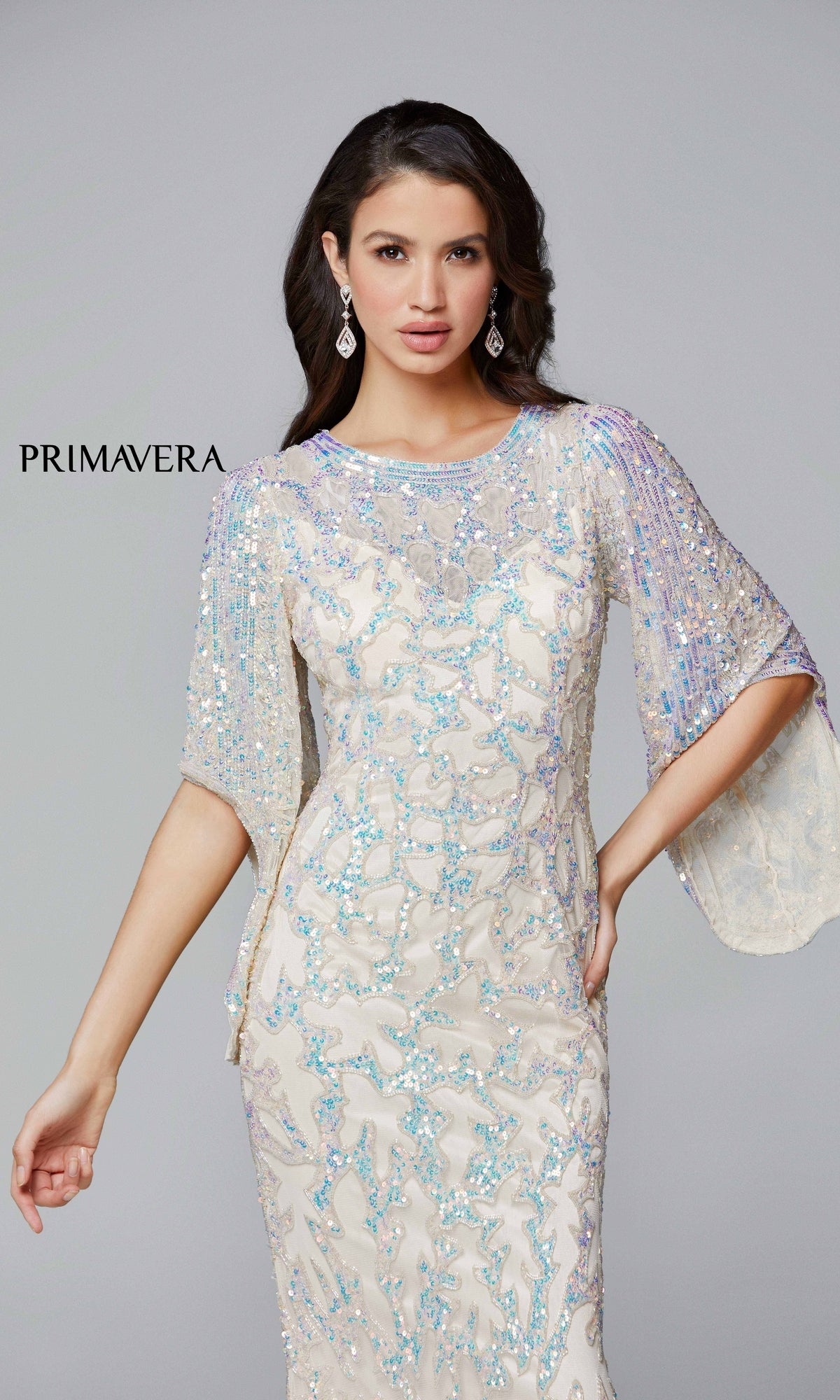 A woman wears the Long Bell Sleeve Beaded Evening Gown 9713, featuring light-colored fabric with intricate blue and silver beading and sheer embellished bell sleeves; 