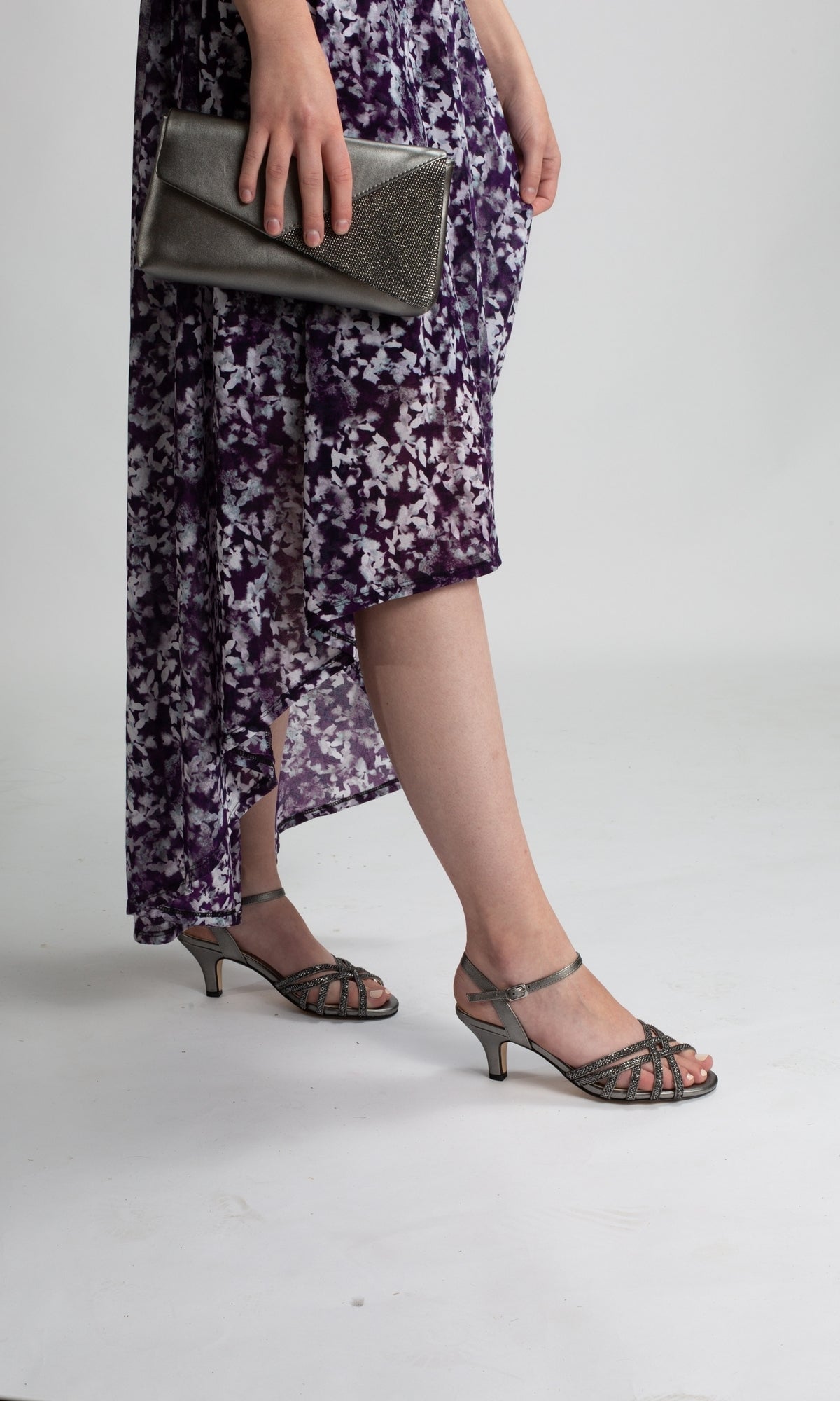 A person stands against a plain background, wearing a purple patterned dress and holding a metallic clutch while showcasing Amara Pewter Open Toe Prom Shoes 4485.