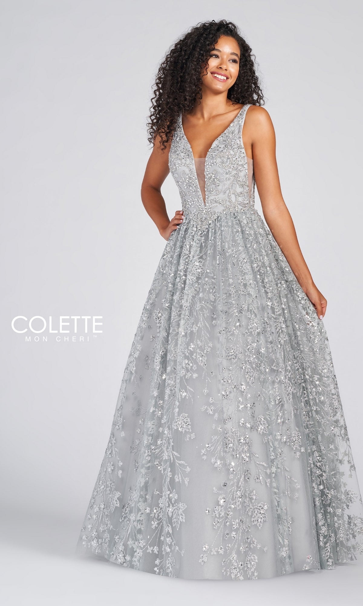 Woman wearing the Colette Designer Prom Sequin & Glitter Ball Gown in silver-grey, featuring a sleeveless design with floral embellishments and a deep V-neckline, standing against a plain background.