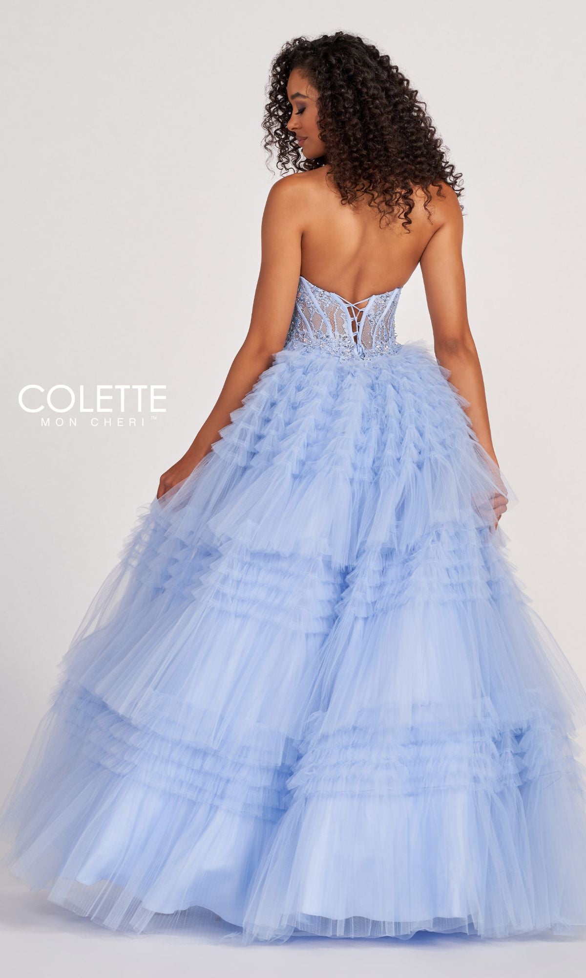 A woman faces away in the Colette CL2017 Strapless Ruffled Prom Ball Gown, featuring a beaded corset bodice and flowing light blue tulle skirt with elegant ruffles.