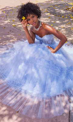 A woman in the Colette CL2017 Strapless Ruffled Prom Ball Gown with a beaded corset bodice sits on a sunlit path, holding a yellow flower and wearing a statement necklace.