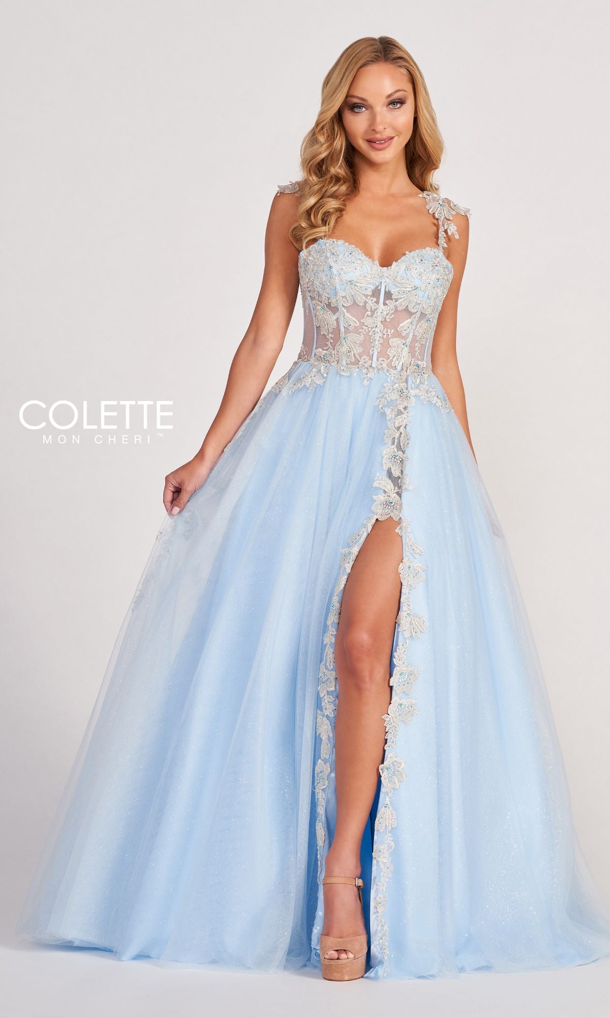 A woman stands against a plain background wearing the Colette CL2020 Glitter-Lace Long A-Line Prom Dress, featuring a light blue color, sheer bodice, floral lace details, and a high front slit.