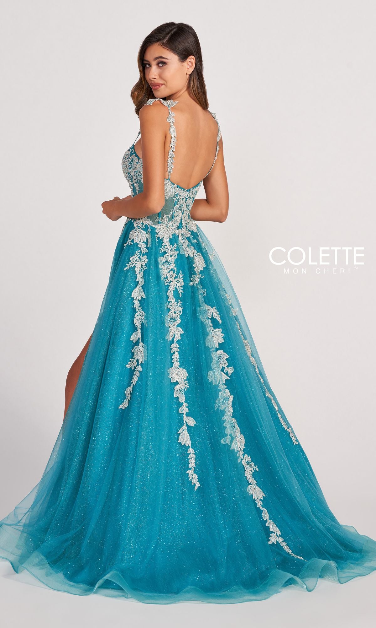A woman in the Colette CL2020 Glitter-Lace Long A-Line Prom Dress stands facing away, glancing over her shoulder. The floor-length turquoise gown features white lace details, an open back, and floral accents for an elegant finish.