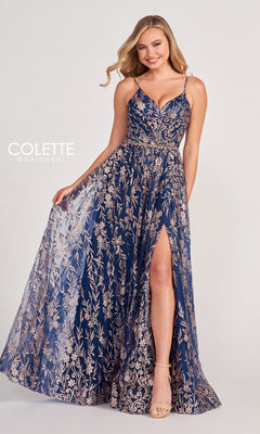 A woman poses and smiles in the Colette CL2022 Strappy A-Line Glitter Prom Dress, featuring a navy blue and gold floral pattern, backless design, and high slit against a plain background.