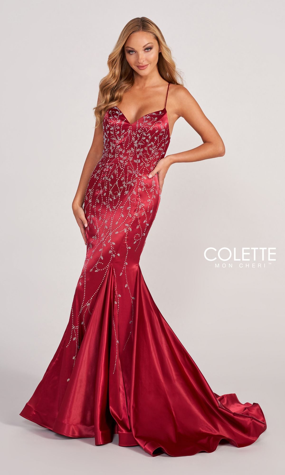 A woman wears the Colette CL2043 Keyhole-Back Long Satin Prom Dress—a fitted, floor-length red gown with a flared hem, thin straps, and silver sequin details—standing against a plain background. 
