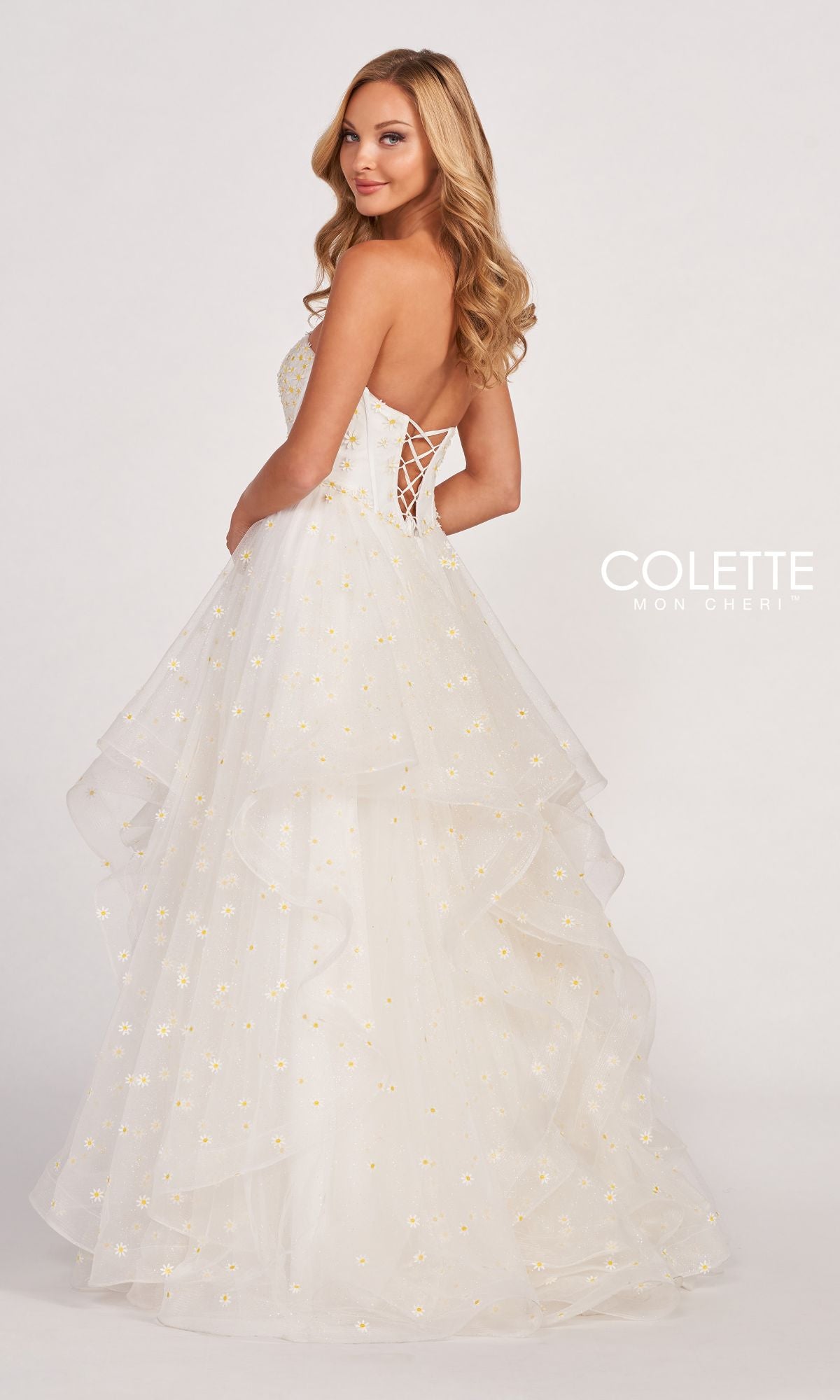 A woman poses in the Colette CL2055 Strapless Daisy Ball Gown, featuring yellow floral details and a lace-up back. She stands against a plain background, glancing over her shoulder.