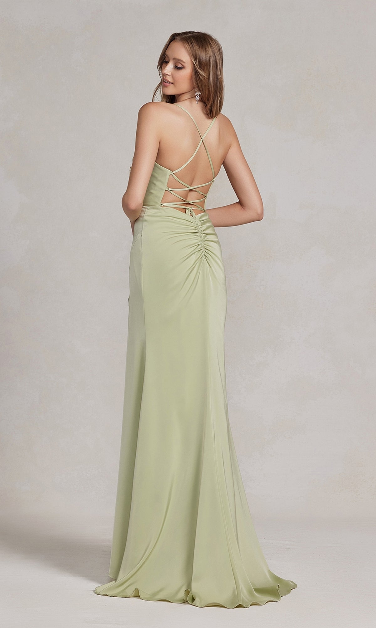 A woman stands facing away in the Cowl-Neck Classic Prom Dress with Side Slit—a light green, floor-length satin gown featuring ruched details and criss-cross straps—an elegant pick for any formal event.