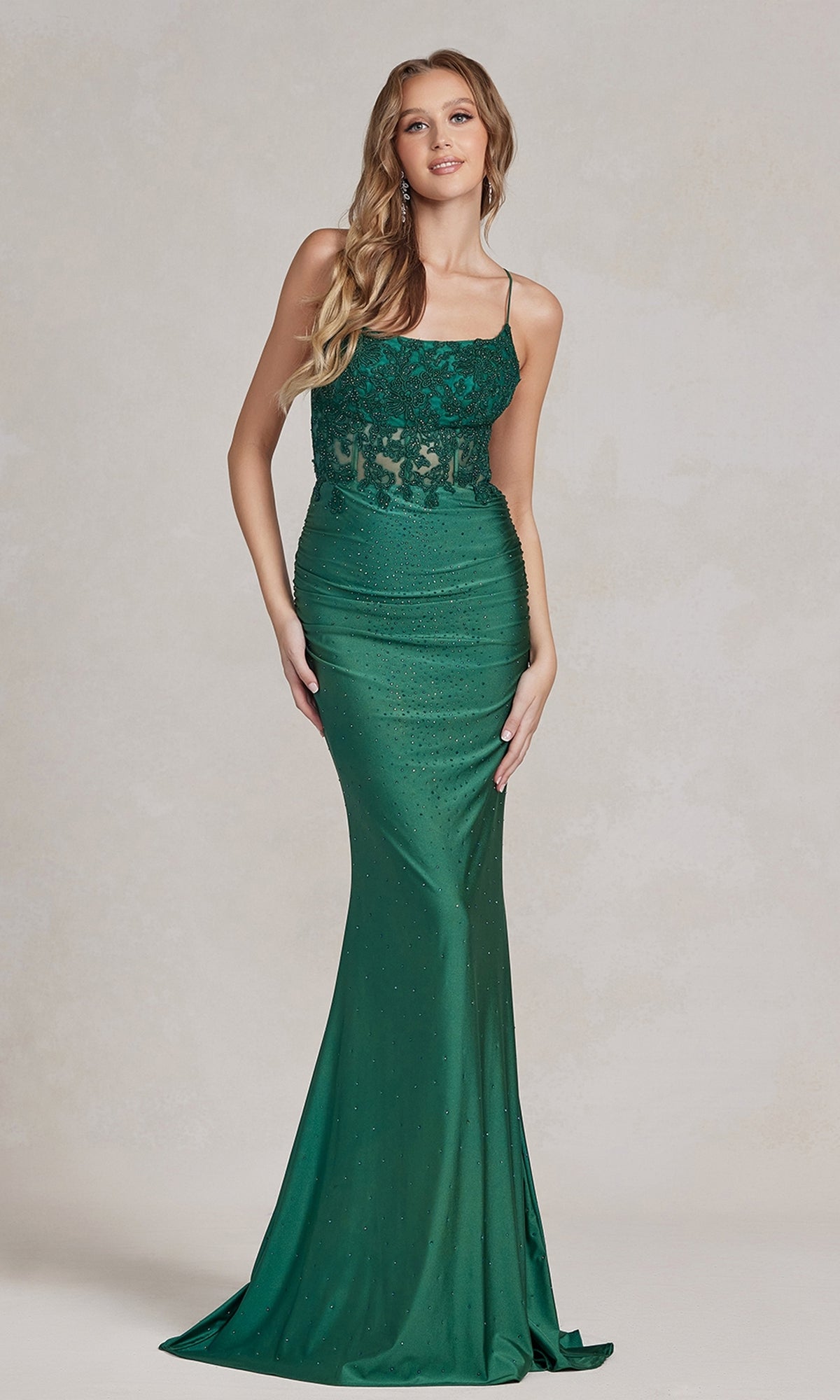 A woman stands wearing a fitted, floor-length green Backless Prom Dress with beaded floral appliques, thin straps, and a lace bodice.