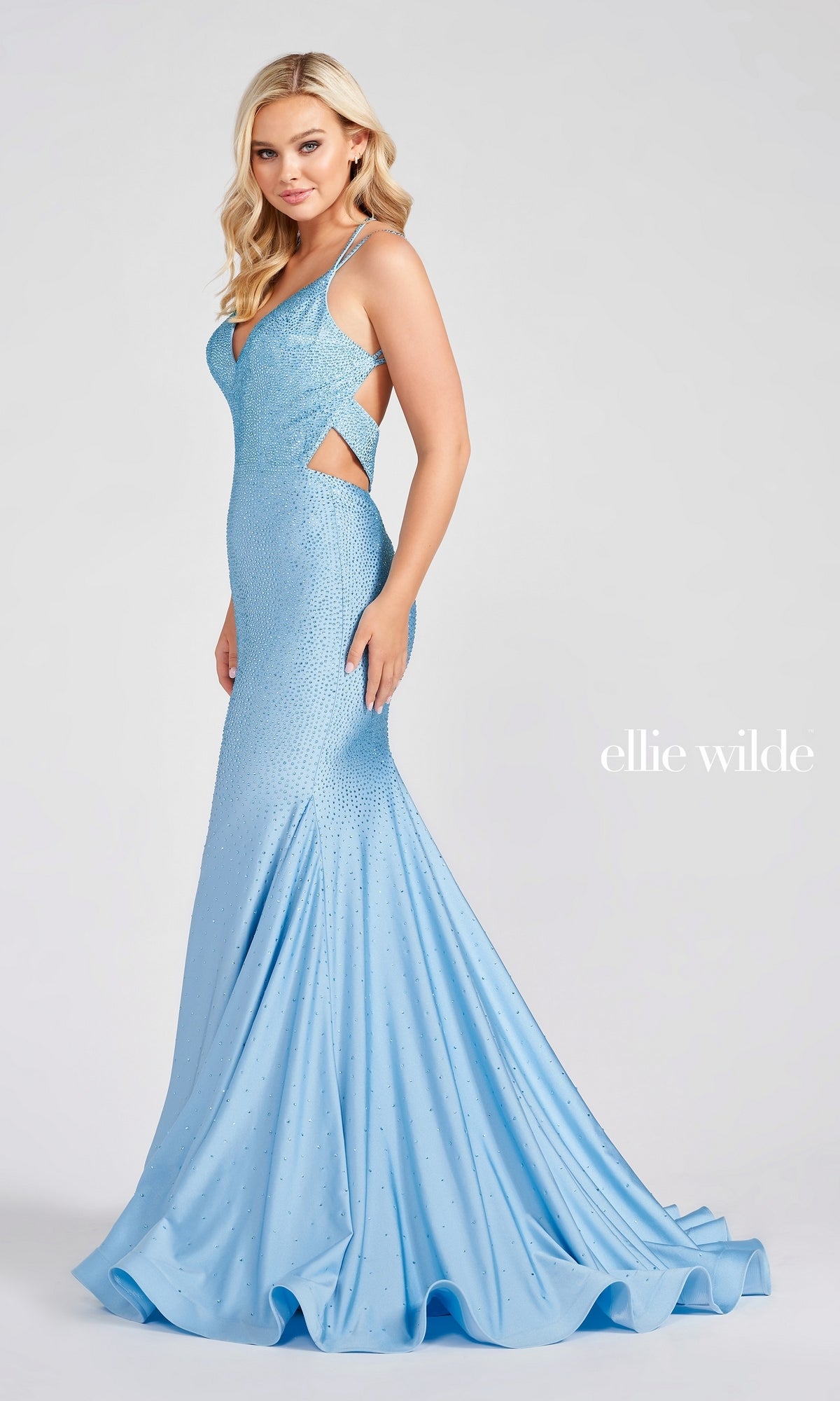 A woman stands against a plain background wearing the Strappy-Back Long Beaded Ellie Wilde Formal Gown—a light blue, sleeveless, floor-length dress with side cutouts, a flared skirt, open back, and elegant bead embellishments.