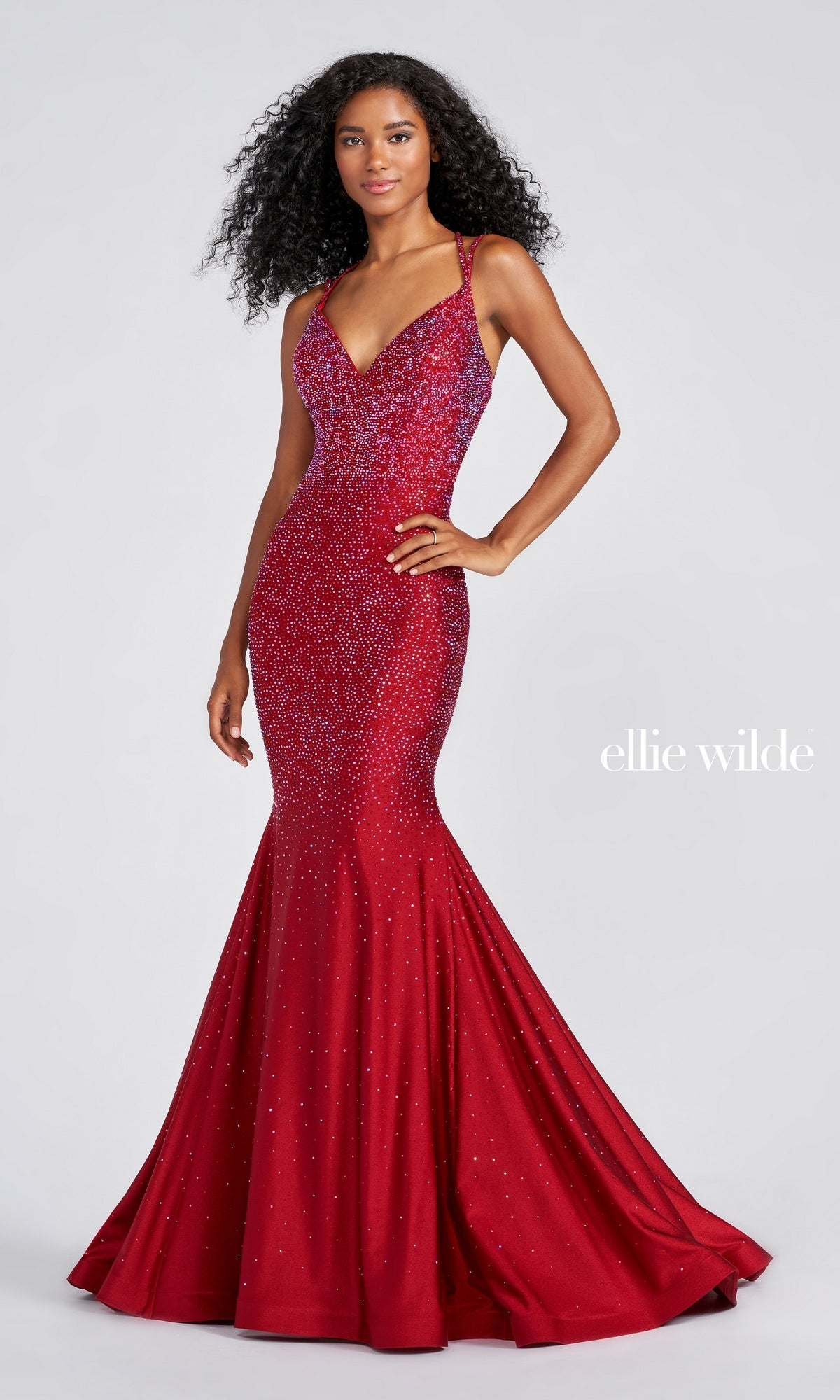 A woman poses in a fitted, sparkly red Strappy-Back Long Beaded Ellie Wilde Formal Gown with thin straps, a subtle gradient effect, and an open-back design. The 